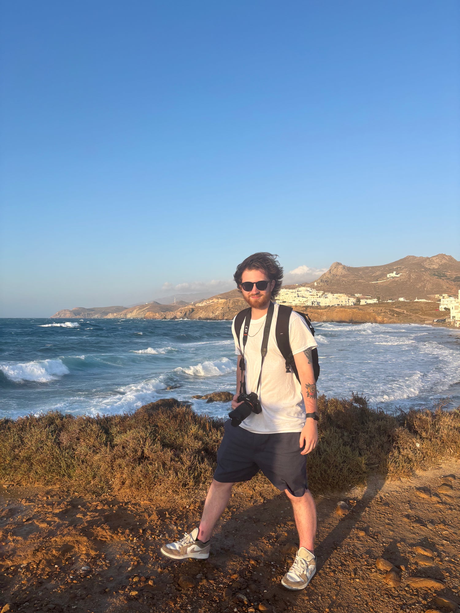 A man wearing a white t-shirt, dark shorts, and sunglasses stands on a rocky cliffside overlooking the ocean with mountains in the background.
